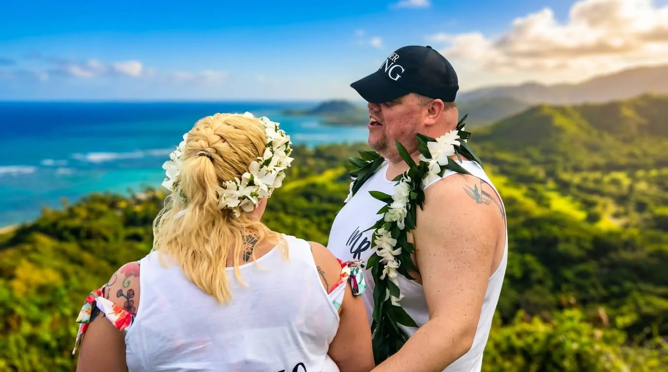 A couple in white clothing and flower leis embracing on a scenic Hawaiian overlook with ocean and mountains in the background.