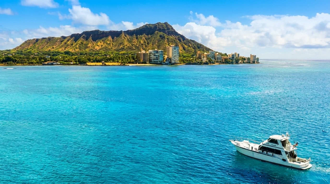 A white boat cruises through brilliant blue ocean waters near Diamond Head crater and Waikiki Beach, with high-rise hotels visible along the shoreline under a partly cloudy sky.