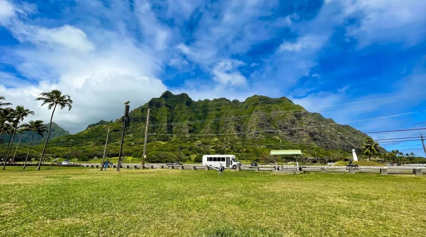 A white tour bus parked on a paved area with lush green mountains in the background, palm trees, and a grassy field in the foreground under a blue sky with white clouds.