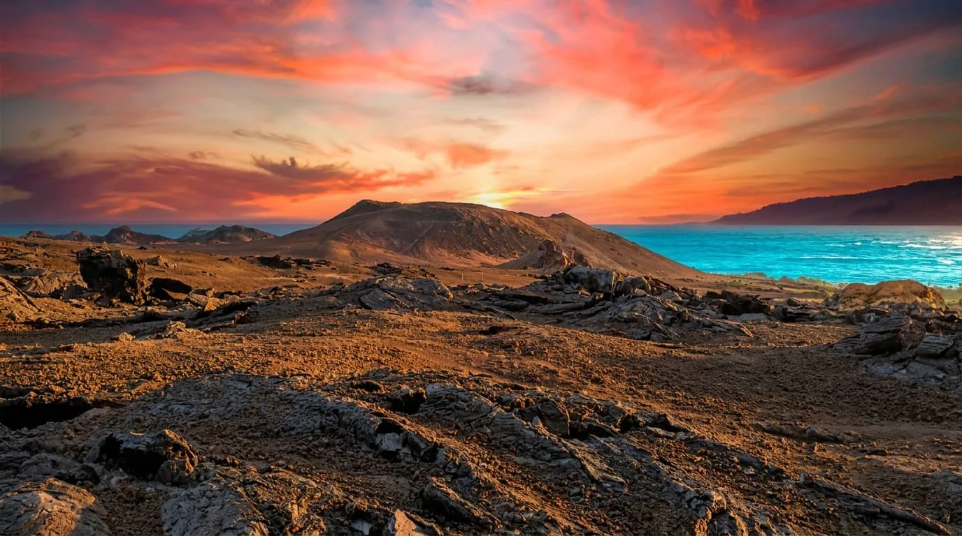 A dramatic sunset over a rugged volcanic coastline with turquoise ocean waters, rocky terrain, and mountains in the distance under a colorful orange and purple sky.