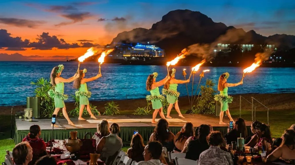 Six female dancers in green traditional Hawaiian costumes performing a fire dance with flaming torches on a stage overlooking the ocean at sunset, with an audience seated at tables watching the performance.