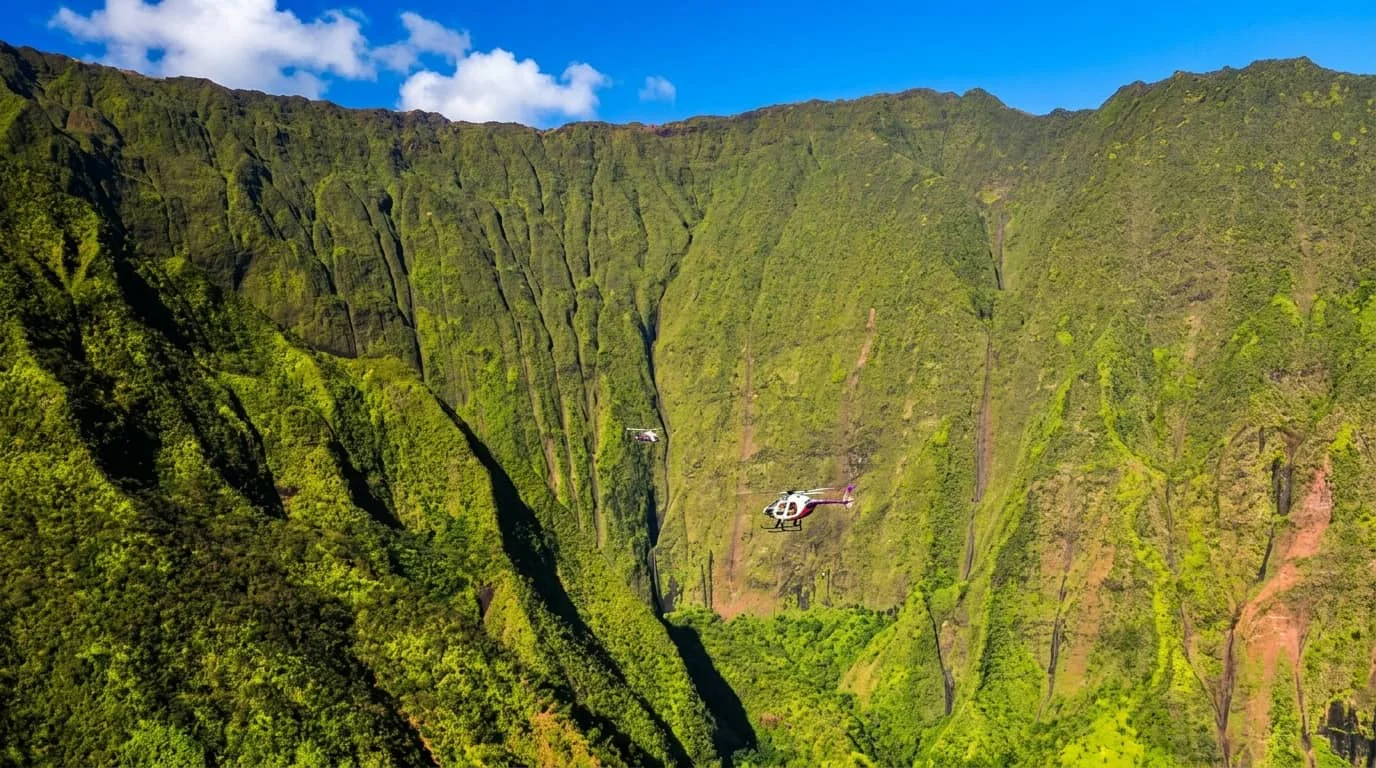 A helicopter flies through a dramatic green mountain valley with steep ridged cliffs and waterfalls under a blue sky with white clouds.