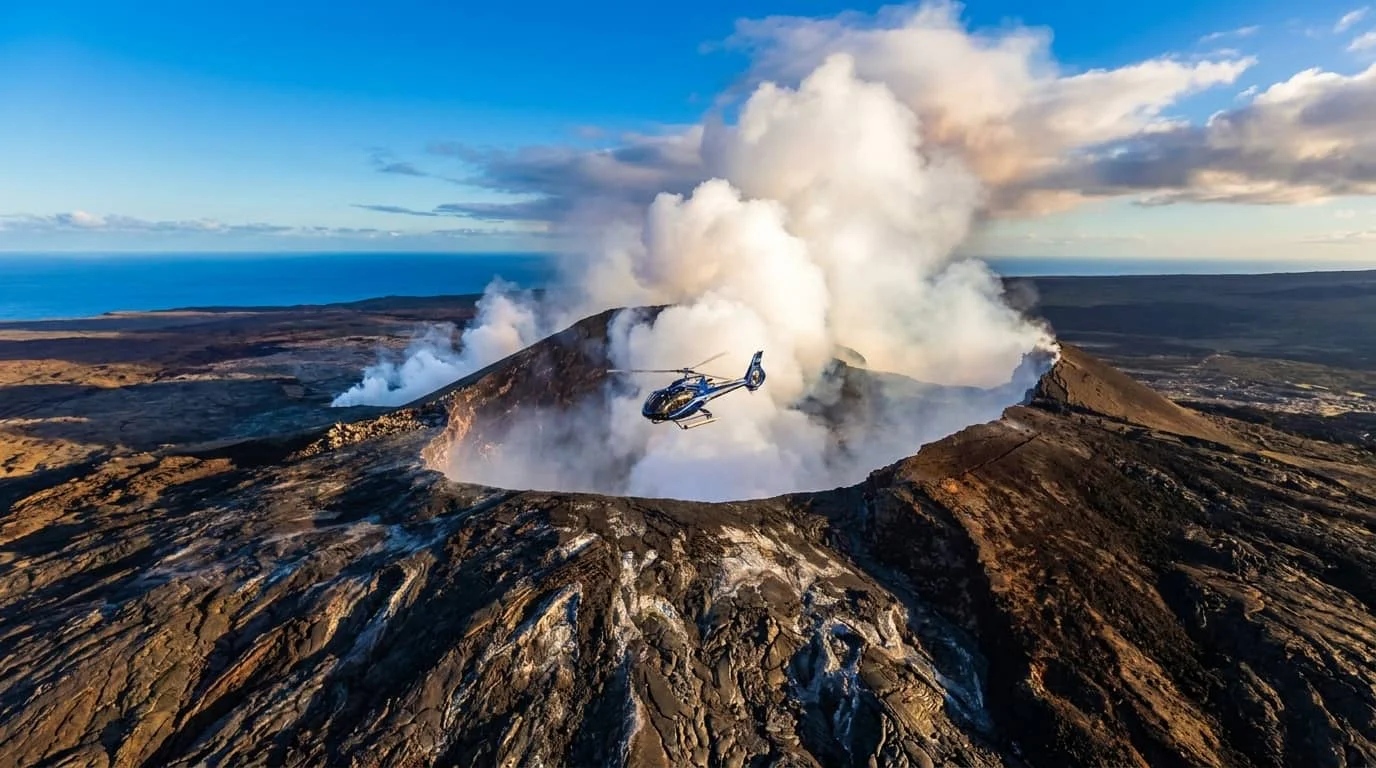 A helicopter flying over an active volcanic crater with billowing white smoke and steam rising from the caldera, showing the dramatic volcanic landscape of Hawaii's Big Island.