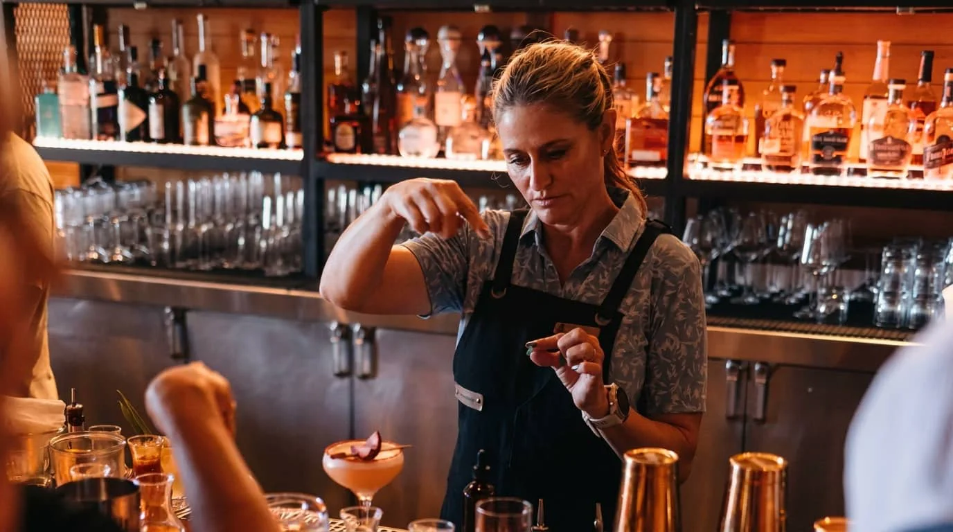 A female bartender in an apron carefully garnishes a cocktail at an upscale bar with shelves of premium spirits and glassware in the background.