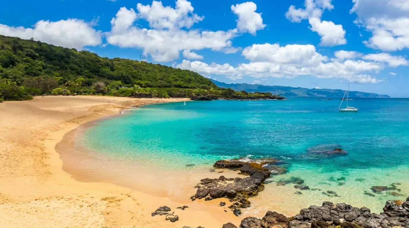 A pristine tropical beach with golden sand, turquoise water, lush green hills, and a white sailboat anchored in the bay under a blue sky with white clouds.