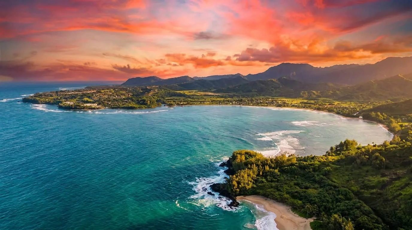 Aerial view of a dramatic Hawaiian coastline at sunset showing turquoise waters, white sand beaches, lush green mountains, and colorful orange and purple clouds in the sky.