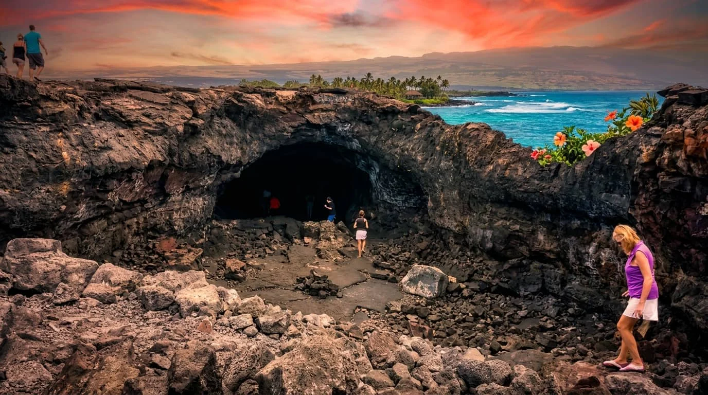 Tourists exploring a large volcanic lava tube cave with dark rocky walls and entrance, with tropical coastline and sunset sky visible in the background.