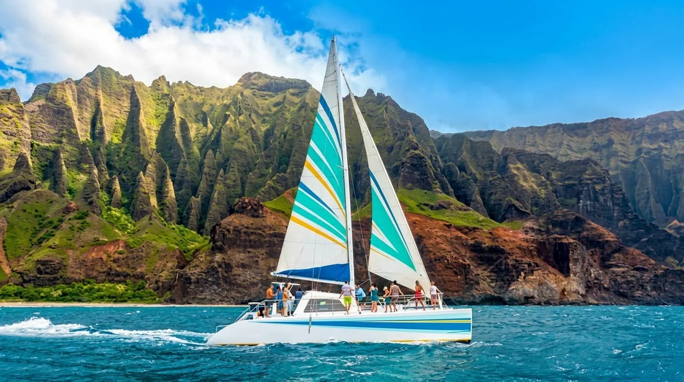 A catamaran sailboat with colorful striped sails cruising through blue ocean waters with dramatic green mountain cliffs in the background.