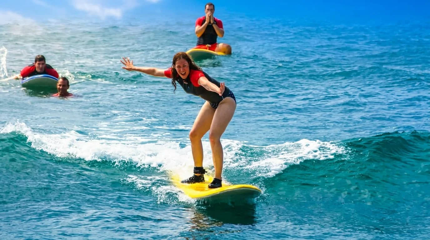 A woman surfing on a yellow surfboard in blue ocean water with other people swimming and floating on boards nearby.