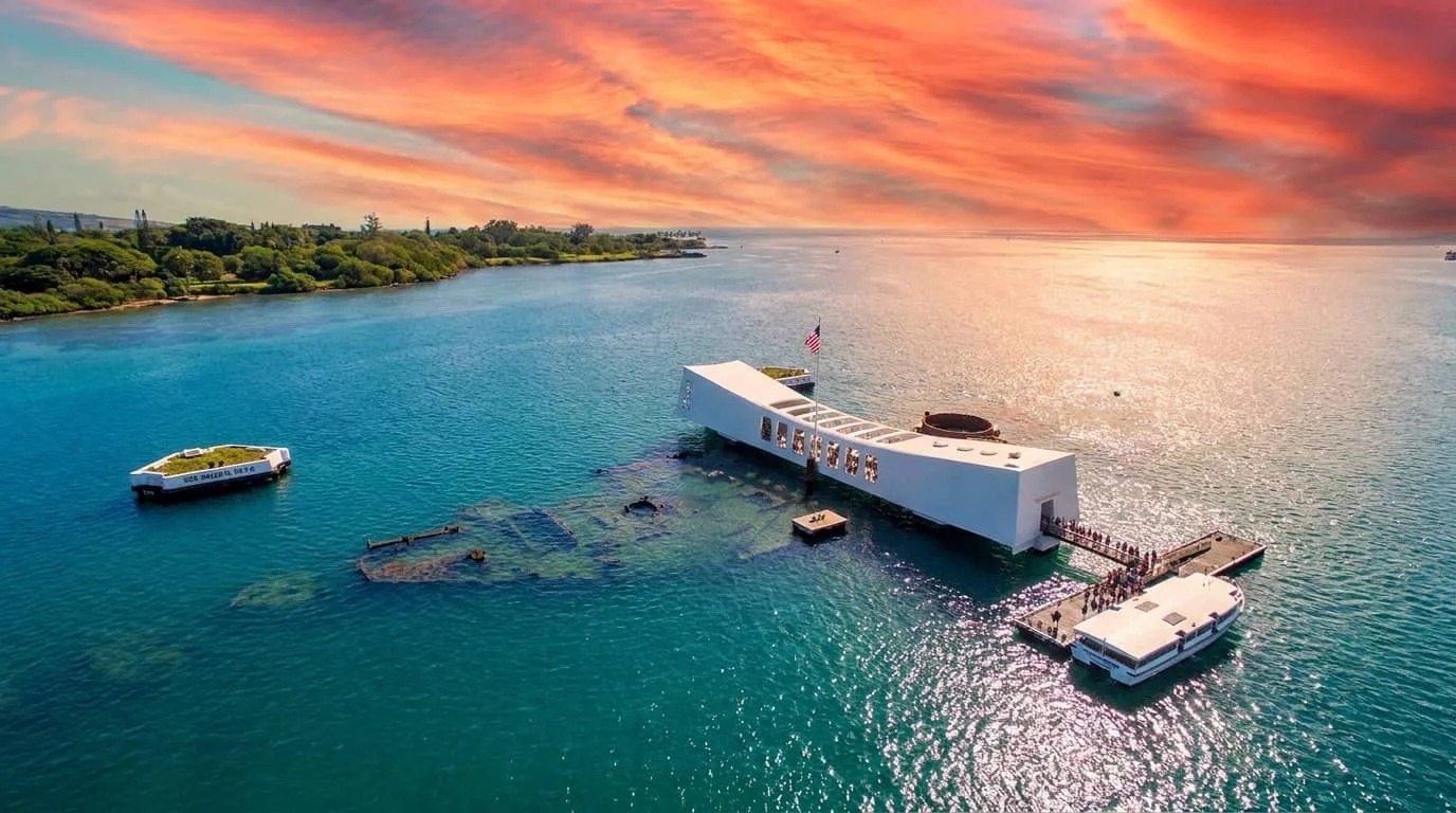 Aerial view of the USS Arizona Memorial at Pearl Harbor with visitors on boats approaching the white memorial structure built over the sunken battleship in blue waters at sunset.