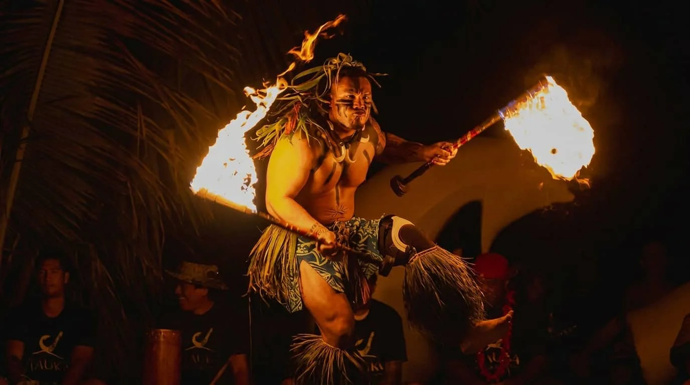 A Polynesian fire dancer performs with flaming torches in traditional costume at an evening luau, with audience members visible in the background under a thatched roof structure.
