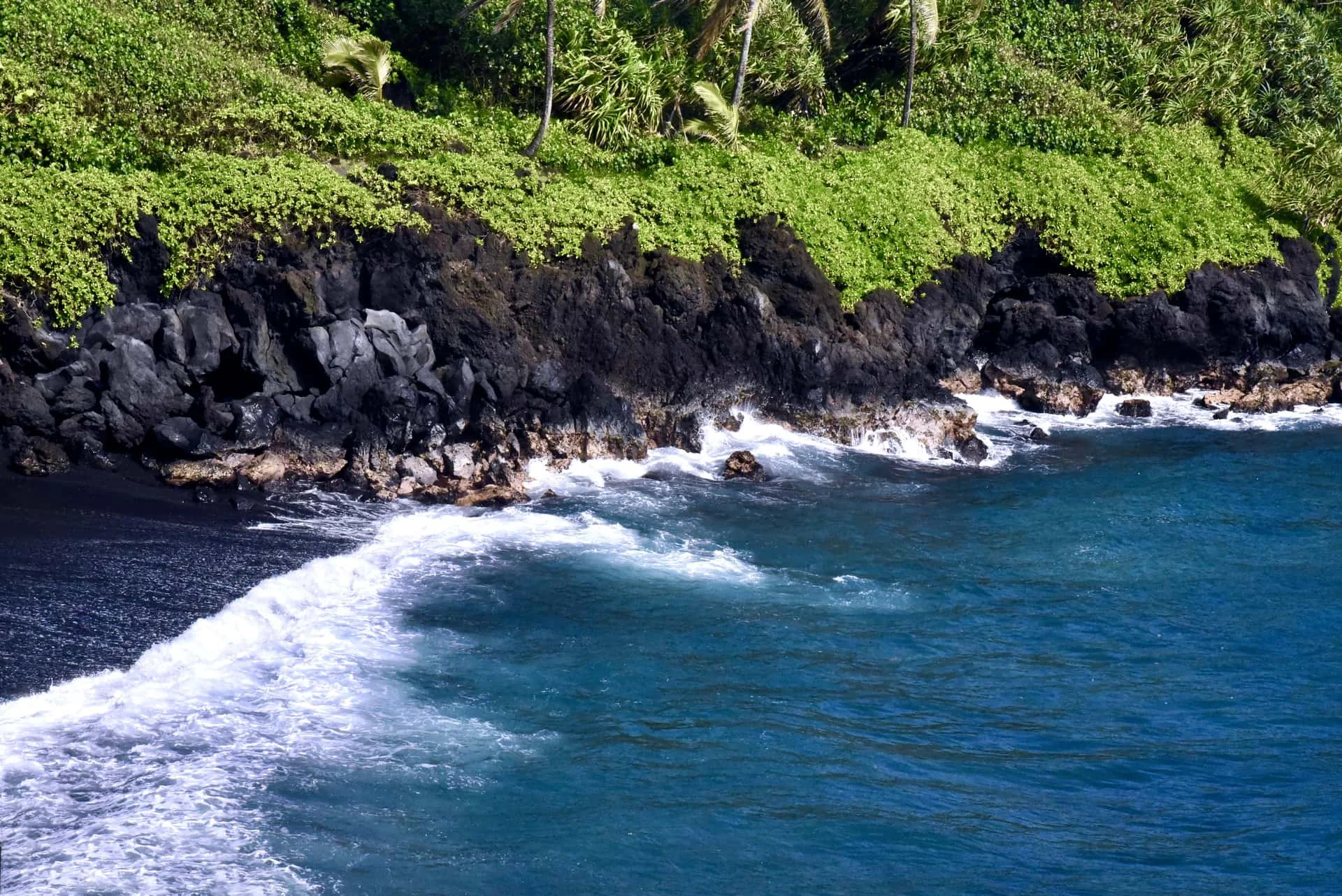 Black sand beach on Maui, Hawaii, with dark volcanic sand meeting turquoise surf and green cliffs