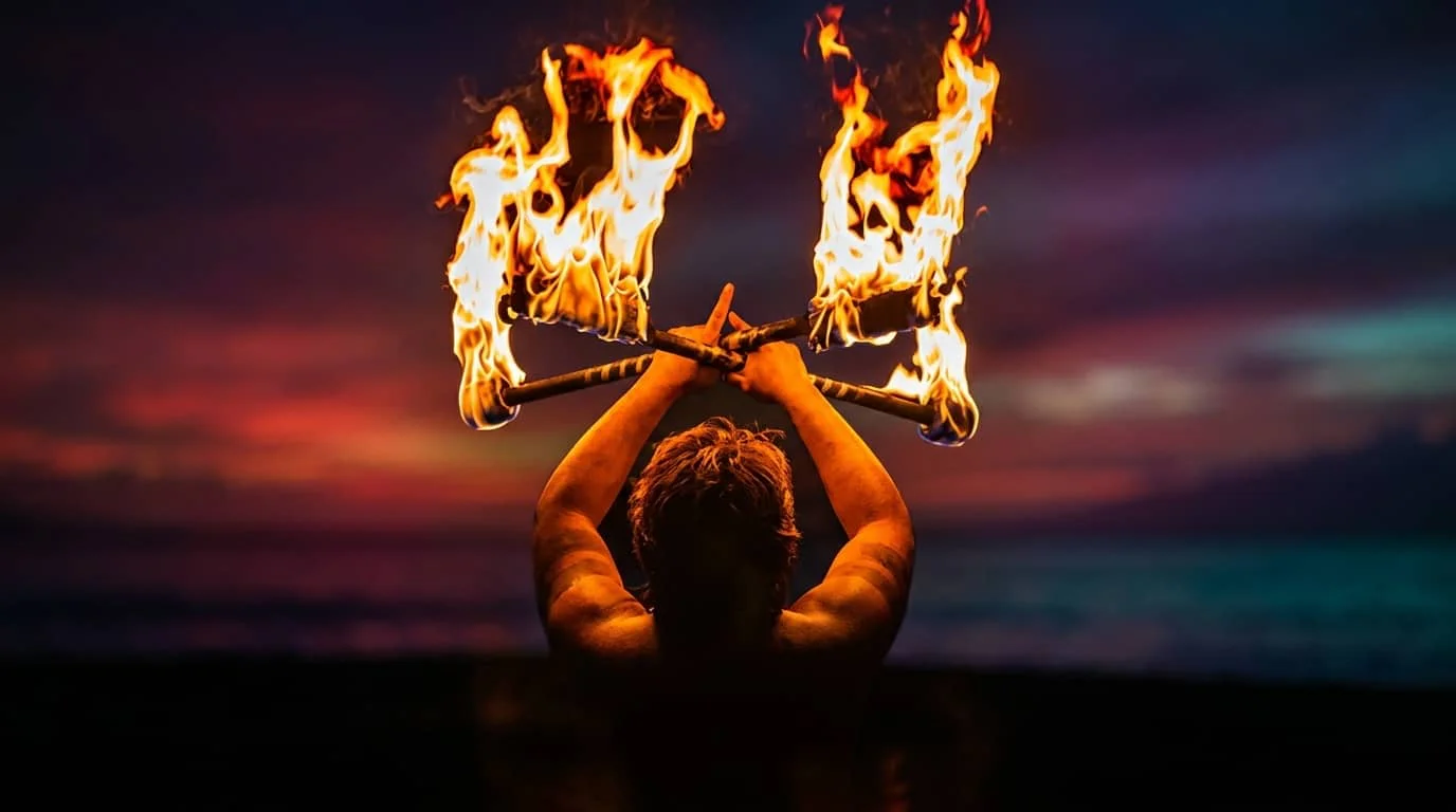 A performer holds two flaming torches overhead against a colorful sunset sky, demonstrating traditional Hawaiian fire dancing.